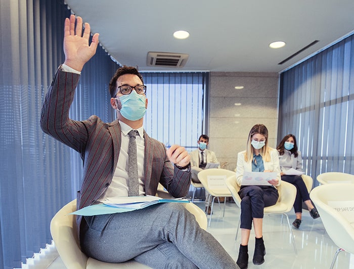 People gathered at desks with masks on with one of them raising their hand up in a social distanced room - Engage Health Care Decision Makers